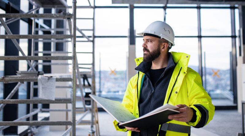 Construction manager in high-visibility jacket and hard hat reviewing site plans inside a building under construction, demonstrating a core role in construction management.