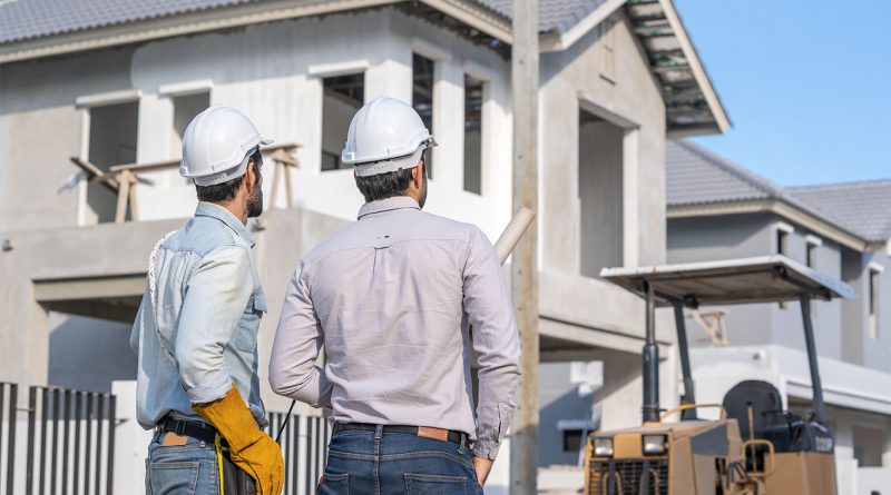 Two construction workers wearing hard hats review blueprints in front of a partially built modern home on a residential construction site.