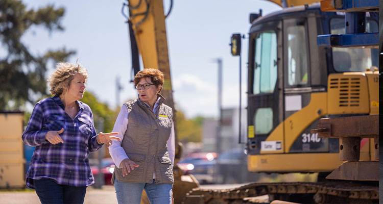 two women standing in front of a large yellow excavator