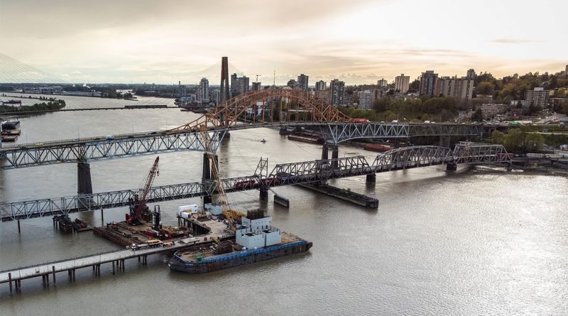 Aerial view of the new Pattullo Bridge tower under construction beside the original bridge over the Fraser River in British Columbia, with cranes, cables, and urban landscape in the background.