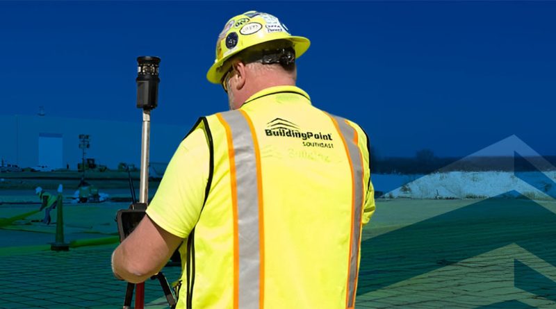 Construction worker wearing a hard hat and high-visibility vest operating a robotic total station on a jobsite, with BuildingPoint Southeast branding visible.