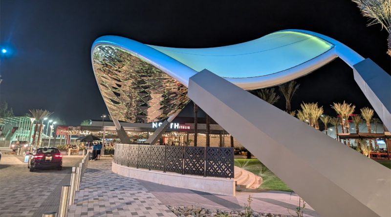 Modern illuminated shade structure at 3A Novus Place in Arizona at night, featuring a sweeping canopy design supported by angled beams, with palm trees, parked cars and a lit commercial entrance in the background.