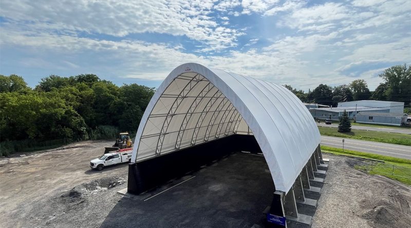 Large white arch-style fabric building with steel frame under construction on a gravel lot, designed for industrial or storage use, with trucks and equipment nearby.